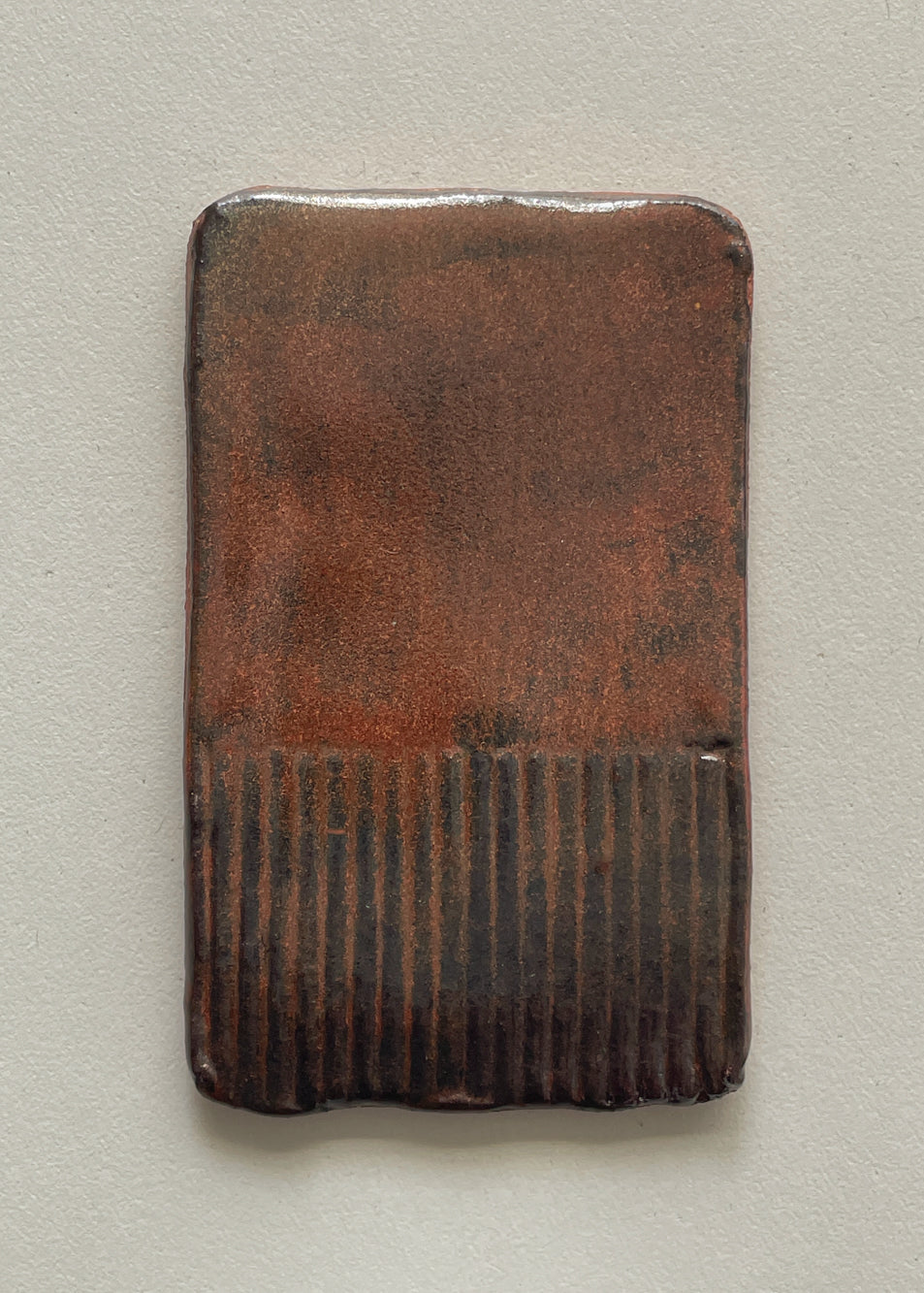A rectangular ceramic tile with rounded corners, featuring a textured reddish-brown glaze on the upper portion and a dark brown ribbed pattern on the lower portion, set against a neutral light gray background.