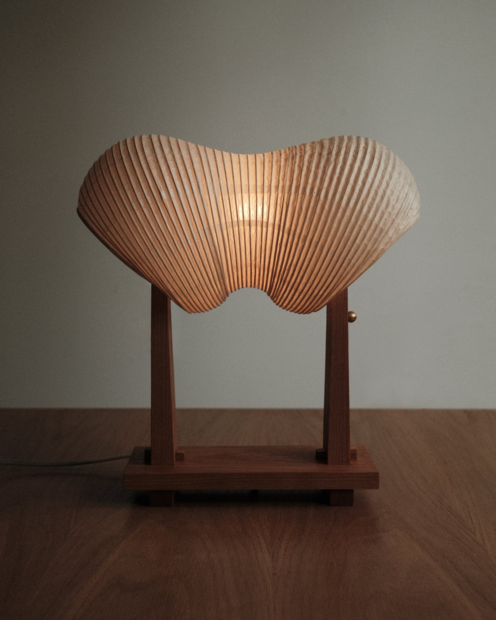 A straight-on shot of Kawabi's Souvenir Table Lamp made of Japanese washi and American Cherry, gently glowing in a dim space.